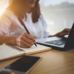 Person writing in notebook using laptop nearby