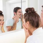 A couple smiling at their reflections in a bathroom mirror while applying skincare products