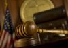 A wooden gavel resting on a desk in a courtroom with an American flag in the background