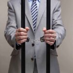 Businessman in a suit holding prison bars with handcuffs on his wrists