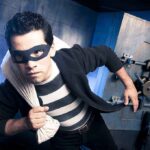 A masked thief in a striped shirt holding a bag while emerging from a bank vault