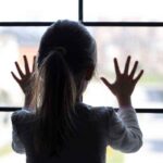 A child with long hair presses their hands against a window, looking outside