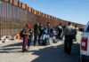 Group of people standing by a border fence with officers and vehicles