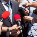 Person in suit surrounded by microphones during media interview