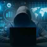 Hooded figure using a laptop in a dark room with data graphics