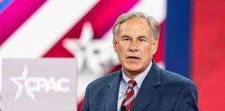 Greg Abbott speaking on stage in a blue suit with a red striped tie