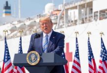 Donald Trump speaking at a podium with American flags and a ship in the background