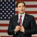 Rubio Schools Media on Trump’s White House Ban man in suit speaking with microphone before American flag