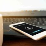 Smartphone with voice assistant on screen beside laptop and coffee mug