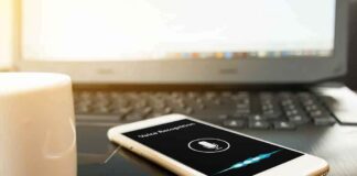 Smartphone with voice assistant on screen beside laptop and coffee mug