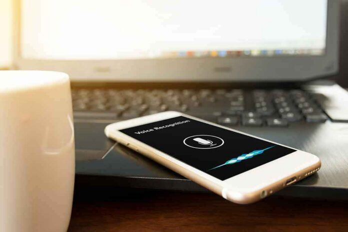 Smartphone with voice assistant on screen beside laptop and coffee mug