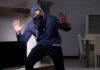 Person in hoodie and face covering raising hands indoors