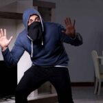 Thief Makes Hilarious Blunder During Home Burglary Person in hoodie and face covering raising hands indoors