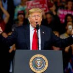 Donald Trump speaking at a rally podium with arms raised