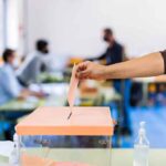 Dems Take Early Edge In Mail-in- Ballots Hand casting ballot into a box at a polling station