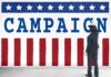 El-Sayed Fires Staffer Who Revealed Funding Bombshell Man in suit writing on large campaign banner with stars and stripes